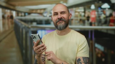 Sakallı bir adam modern bir alışveriş merkezinde akıllı telefon kullanıyor. Şehir yaşamını geniş ışıklandırmalı, hareketli bir ev ortamında sergiliyor..