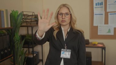 Ofiste duran, gözlük takan ve arkaplan çizelgesi olan siyah takım elbiseli bir kadın, kapalı alanda profesyonel bir ortam izlenimi veriyor..