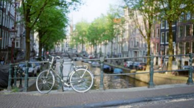 Bisiklet, arka planda parlak güneş ışığı altında bulanık binalar, ağaçlar ve su bulunan bir kanal köprüsüne odaklanmış vaziyette duruyor..