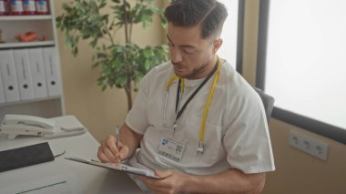 Sakallı genç bir doktor, parlak bir klinik odasında notlar yazıyor. Steteskop ve beyaz önlük giyiyor. Evrak işleri ve organizasyonla dolu profesyonel bir tıbbi ortama işaret ediyor..