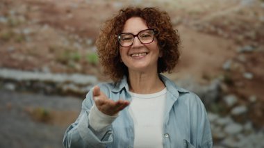 İspanyol orta yaşlı bir kadın şehir sokaklarında dostça bir gülümsemeyle elini uzatıyor, gözlük takıyor ve günlük giysiler giyiyor, şehir manzarası ortamında etkileşime girmeye davet ediyor..