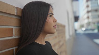 Uzun saçlı genç bir kadın şehir sokaklarında duvara yaslanıyor, sakin bir ifade sergiliyor ve açık havada düşünceli davranışlar sergiliyor..