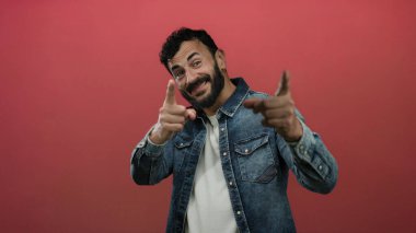 Sakallı İspanyol bir adam gülümsüyor ve kırmızı arka planda kot ceket giyiyor..