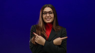 Gözlüklü ve siyah takım elbiseli İspanyol kadın canlı mavi arka plana karşı güvenle yanlamasına bakıyor, yönünü ve kararını odaklanmış bir ifadeyle gösteriyor..