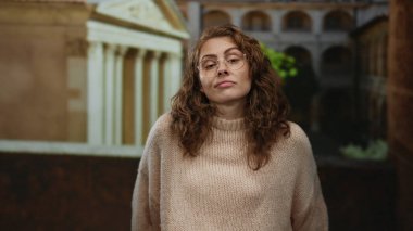 Eski bir üniversitede gözlüklü ve kıvırcık saçlı bir süveter giyen bir kadın klasik mimariyle çevrili düşünceli bir ifade sergiliyor..