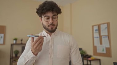 Ofisteki Latin kökenli genç adam sesli mesaj kaydediyor. Odaklanmış ifadesi ve günlük kıyafetleri olan akıllı telefon kullanıyor. Etrafı notlar ve ev ortamındaki bitkilerle çevrili.