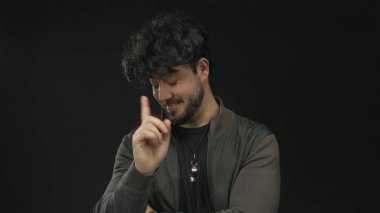 Sakallı ve kıvırcık saçlı genç adam siyah bir arkaplana karşı jest yapıyor, koyu renk ceketli ve gündelik kıyafetli oyuncu ve düşünceli bir tavır sergiliyor..