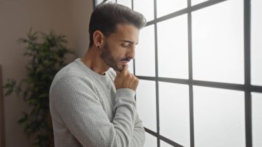 İçerde rahat bir ortamda genç bir İspanyol adam, büyük bir pencereden dışarı dikkatlice bakıyor, şık bir apartman dairesinde düşünce ve iç gözlem atmosferi yayıyor..