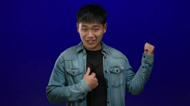 Kot giyen genç adam, izole edilmiş mavi arka plana karşı özgüvenle el sallıyor.