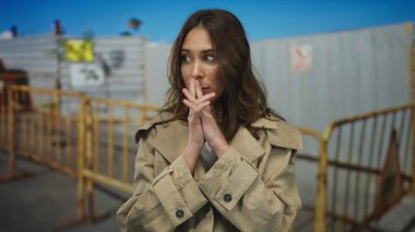 Genç bir kadın arka planda bariyerler olan bir inşaat alanında duruyor, bir trençkot giyiyor ve açık hava manzarasına karşı düşüncelerini dile getiriyor..