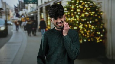 Şehir sokağında genç bir adam tatil sezonunda yüzü ağrıyan bir diş ağrısı çekiyor ve arka planda bir Noel ağacı var..