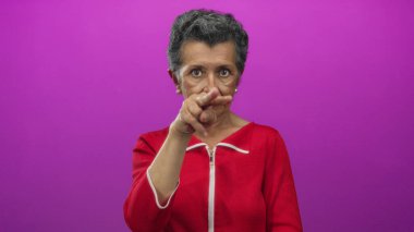 Kırmızı elbiseli yaşlı bir kadın canlı pembe bir arka plana karşı ihtiyatlı bir jest yapıyor..