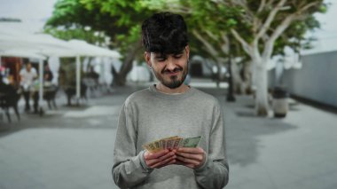 Şehir caddesinde Norveçli bir kronu elinde tutan bir adam yaya yolu boyunca uzanan ağaçlarla, şehirde sıradan bir açık hava manzarası tasvir ediyor..