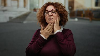 İspanyol, orta yaşlı, kıvırcık saçlı bir kadın şehir caddesinde duruyor, gözlük takıyor, yüzüne krem sürüyor..