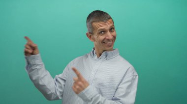 Stüdyo ortamında yeşil bir duvara gülümseyen ve işaret eden bir adam mutluluğu, pozitifliği ve açık mavi gömlekli günlük kıyafetleri vurguluyor..