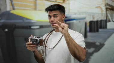 Şehir sokağındaki İspanyol asıllı genç adam elinde antika bir kamera tutarken parmaklarıyla şakacı jestler yapıyor ve canlı şehir sahnesini yakalıyor..