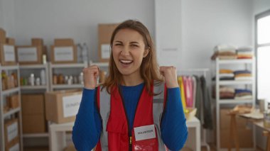 İç mekan hayır kurumunda gönüllü olarak gülümseyen genç bir kadın. Kırmızı yelek giyiyor. Etrafı düzenli kutular, malzeme ve kıyafetlerle dolu..