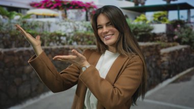 Genç İspanyol kadın gülümsüyor ve elleri ile canlı bir şehir caddesinde etrafı renkli çiçekler ve açık hava dekorlarıyla çevrili öğleden sonra ışığında şık bir ceket giyiyor..