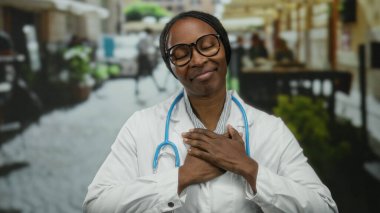 Gözleri kapalı, gözlüklü ve laboratuvar önlüklü bir kadın canlı bir sokak ortamında, boynuna steteskopla şükranlarını sunuyor..