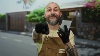 Sakallı bir adam, şehir sokaklarında defter tutuyor, eldiven ve önlük takıyor, motosikletli bulanık bir arka planı var..