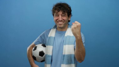 İspanyol atkı takan ve elinde futbol topu tutan adam mavi arka planda neşeli bir ifadeyle spor hevesini ve zaferi sembolize ediyor..