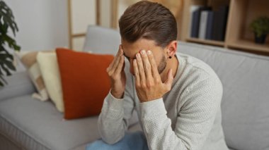 Kanepede oturan genç bir adam modern bir apartman dairesinde yüzünü örterek stres ve tefekkür gösteriyor..