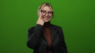 Siyah ceketli, yeşil arka planda gözlüklü, parlak renklerde bir stüdyo ortamında kendine güveni ve pozitifliği gösteren bir kadın..