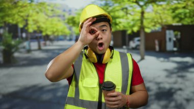 İnşaat işçisi, elinde kahve, çelik yelek ve kulaklık ile şehir merkezinde güvenli bir şekilde etrafa bakıyor.