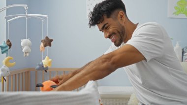 Evdeki yatak odasındaki beşikte gülümseyen genç adam, bebek oyuncaklarıyla meşgul, sıcaklığını, neşesini ve aile ortamında ebeveynlik yapıyor..