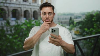 Sakallı genç adam Roma kolezyumunun yanında dikilirken akıllı telefona şaşırdı, arka planda yeşillik olan açık hava sahnesini gün ışığında çekti..