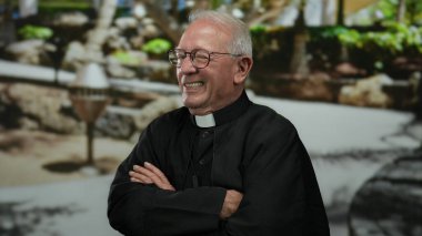 Sokak ortamında gülümseyen yaşlı bir rahip papaz tasması takıyor ve kolları çaprazlanmış gözlükler takıyor..