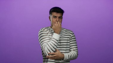 İspanyol asıllı genç, çizgili gömlek içinde, izole edilmiş mor arka planda endişeli görünüyor..