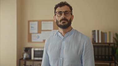 Gözlüklü ve sakallı bir adam düşünceli bir şekilde ofiste duruyor. Etrafı ilan panosu, mikrodalga fırın, kitaplar, tarafsız duvarlara dayanan kahve modern çalışma alanlarıyla çevrili..