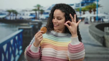 Gezintide elinde kurabiyeyle gülümseyen bir kadın, renkli bir kazak giyiyor, canlı şehir manzarası arasında hareketli bir hareket sergiliyor, neşeli bir açık hava anını yakalıyor..