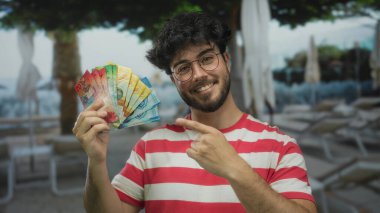 Sakallı genç bir adam, İsviçre frangını bir tatil köyünde havuz kenarında tutuyor. Rahatça gülümsüyor..