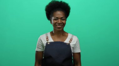 Önlük takan kadın parlak yeşil arka plana karşı duruyor kendine güven ve duruş yayıyor..