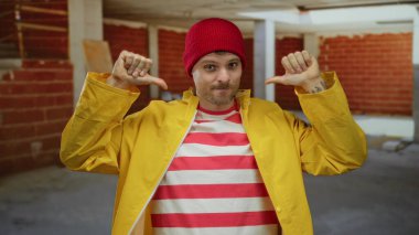 İnşaat alanında sarı yağmurluk ve kırmızı bere giyen kendine güvenen, çizgili gömlekli çekici bir adam..