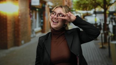 Bir kadın gülümsüyor ve şehir sokaklarında barış işareti yapıyor, gözlük ve koyu renk bir ceket giyiyor, gündüzleri güven ve mutluluk saçıyor..