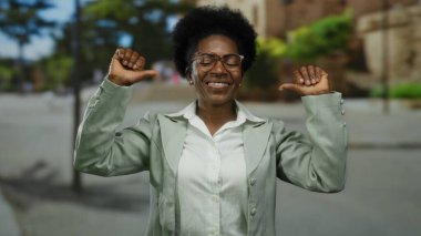 Gözlüklü bir kadın, yeşil bir caddede kendine güveni tam bir şekilde kendini gösteriyor. Açık renk bir ceket giyiyor ve hareketleriyle neşe saçıyor..