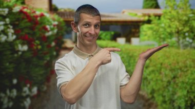 Açık havada gülümseyen bir adam iyi bir işaret ve parlak bir sokakta açık bir avuç içi ile, güneşli bir günde renkli çiçekler ve yemyeşil ile çevrili..