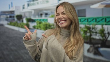 Balıkçı yaka kazak giyen sarışın kadın şehir sokaklarında gülümseyen parmağıyla işaret ediyor iyimserlik rehber neşe güveni.