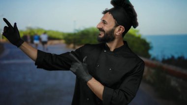 Siyah üniformalı İspanyol aşçı deniz kenarında gülerken arka planda yemyeşil ve mavi suyla canlı bir açık hava manzarası olduğunu gösteriyor..