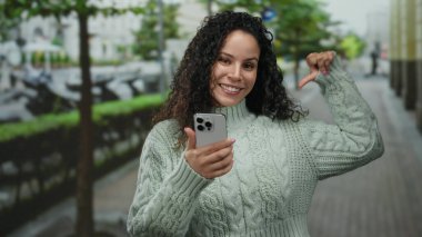 Genç İspanyol kadın kendini göstererek gülümsüyor ve açık bir sokak ortamında elinde bir akıllı telefon tutuyor, bulanık bir şehir arka planına karşı örülü bir süveter giyiyor..