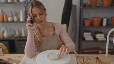 Önlüklü kadın stüdyoda kil ve tekerleği nazikçe şekillendirmek için elleri kullanır; yaratıcılık farkındalığı odak noktası.