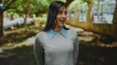 Arka planda ağaçlarla parkta dikilen bir kadın, dışarıda güneşli bir günde merak ve sürpriz sergiliyor..