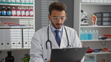 Hastane odasındaki erkek doktor tıbbi malzeme ve ekipmanlarla çevrili bir panoyu okuyarak profesyonel bir sağlık hizmetleri ortamını vurguluyor..