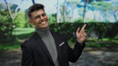 Açık hava parkında gülümseyen İspanyol bir adam gözlüklü ve günlük ceketli, kendine güvenen ve doğal çevrede çekici bir tavır sergiliyor.