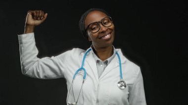 Beyaz önlüklü, steteskoplu bir kadın güçlü ve profesyonelliğini gösteren koyu arkaplana karşı yumruğunu kaldırıyor.
