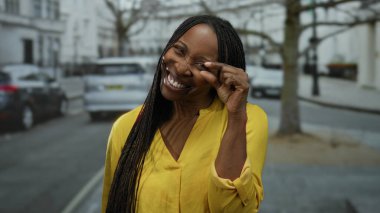 Kıvırcık saçlı gülümseyen kadın şehir sokaklarında jest yapıyor. Bulanık şehir arkaplanıyla neşeli ve neşeli bir atmosfer sergiliyor..