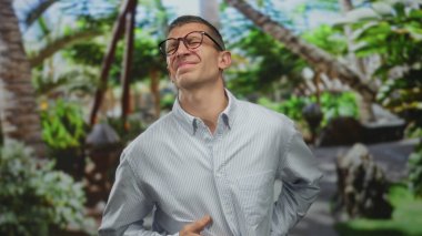 Parkta gözlüklü ve mavi gömlekli beyaz bir adam yeşil bir arka planda karın ağrısı çekiyor. Rahatsızlık veriyor ve doğa düzenini kuruyor..